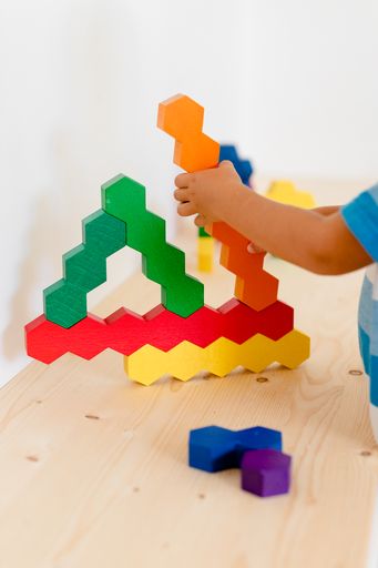 Kind spielt mit Honeycomb Holzbausteinen auf dem Boden. Es sind die Bausteine zu sehen, sowie der Arm des Kindes und seine Hand, die einen orangen Baustein zum Bauwerk hinzufügt. Ganz rechts ist ein kleines Stück seines Beins zu sehen