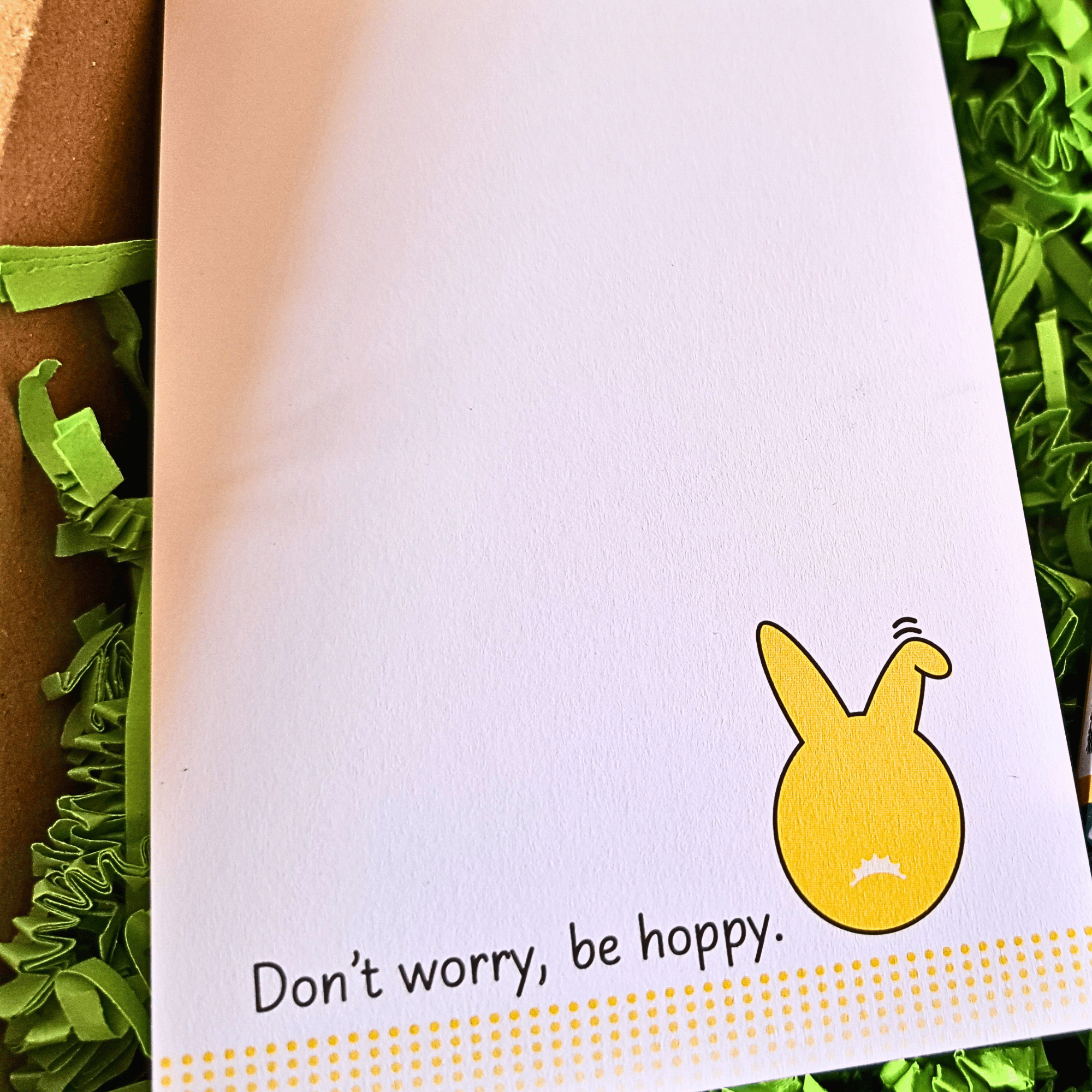 Detailansicht der unteren Ecke des Notizblocks. Die gelbe Hasenrückseite und den Schriftzug "Don’t worry, be hoppy." sind deutlich auf weißem Papier zu sehen. Vier gepunktete Linien werden nach unten hin transparenter. Der Block liegt eingebettet in grüne