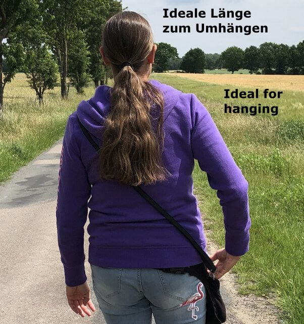 Frau mit lila Kapuzenjacke und Jeans ist von hinten zu sehen. Sie geht einen Feldweg entlang und hat eine Hundeleine quer über die Schulter gehängt.