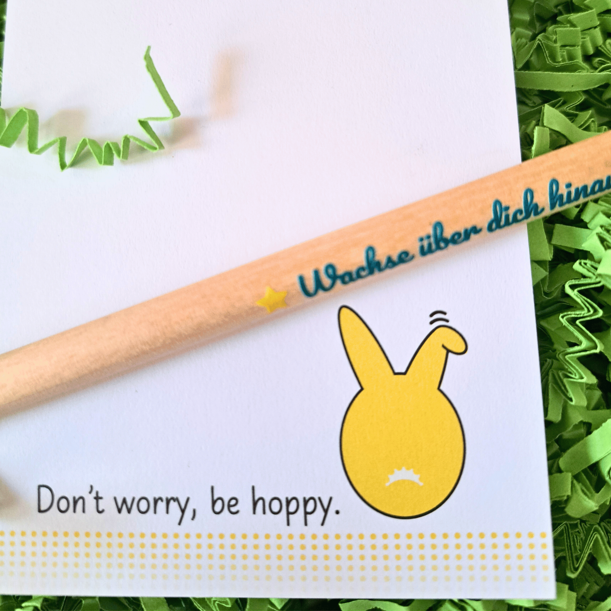 Nahaufnahme: Oster-Notizblock und einpflanzbarer Bleistift Nahaufnahme: Notizblock mit gelbem Hasen und Spruch „Don’t worry, be hoppy.“ Quer darüber ein Bleistift mit dem Text „Wachse über dich hinaus!“. Hintergrund grünes Gras.
