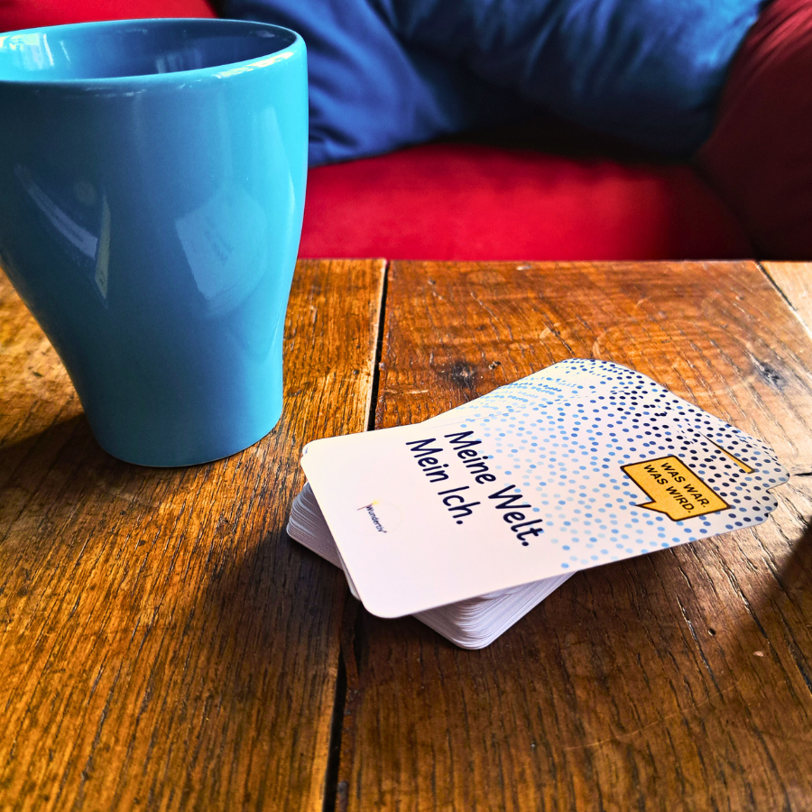 Eine türkis Tasse auf einem Holztisch. Neben der Tasse liegt ein Stapel Gesprächswunder-Karten "Was war. Was wird. Meine Welt. Mein ich.". Im Hintergrund ist ein zu roter Sessel mit türkis Kissen zu sehen.
