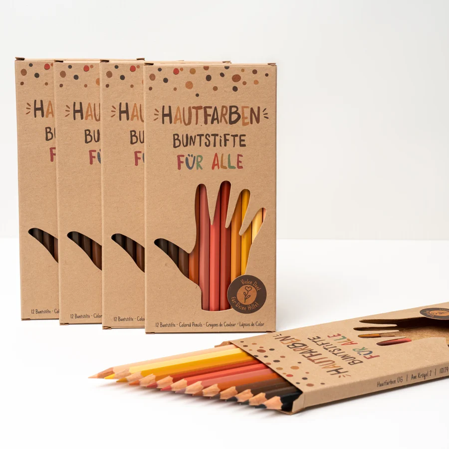 Seitliche Aufnahme Verpackung des Hautfarben-Stifte-Sets mit hervorstehenden Stiften - Hintergrund Stiftverpackungen Hochkant Eine Verpackung der Hautfarben Buntstifte liegt schräg auf einem weißen Hintergrund. Einige Stifte ragen aus der Verpackung heraus und zeigen schräg zum Betrachter. Im Hintergrund stehen vier Verpackungen hochkant