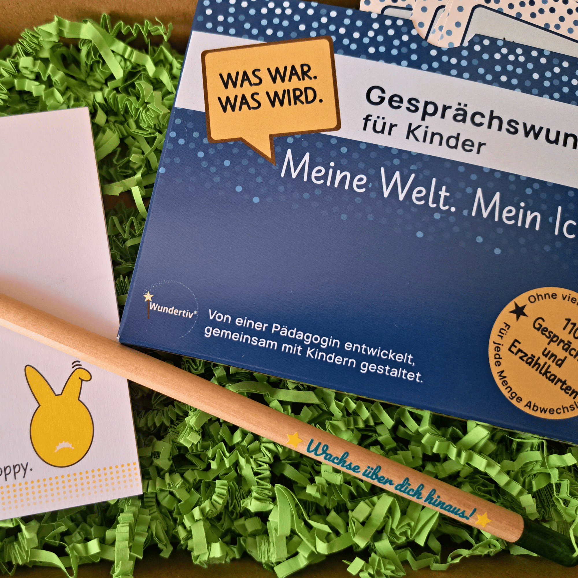 Detailansicht Gesprächswunder Osterspecial mit Oster-Zubehör Nahaufnahme Gesprächswunder Set „Meine Welt. Mein Ich.“ auf grünem Gras mit Samenbleistift „Wachse über dich hinaus!“ und Notizblock-Ecke.