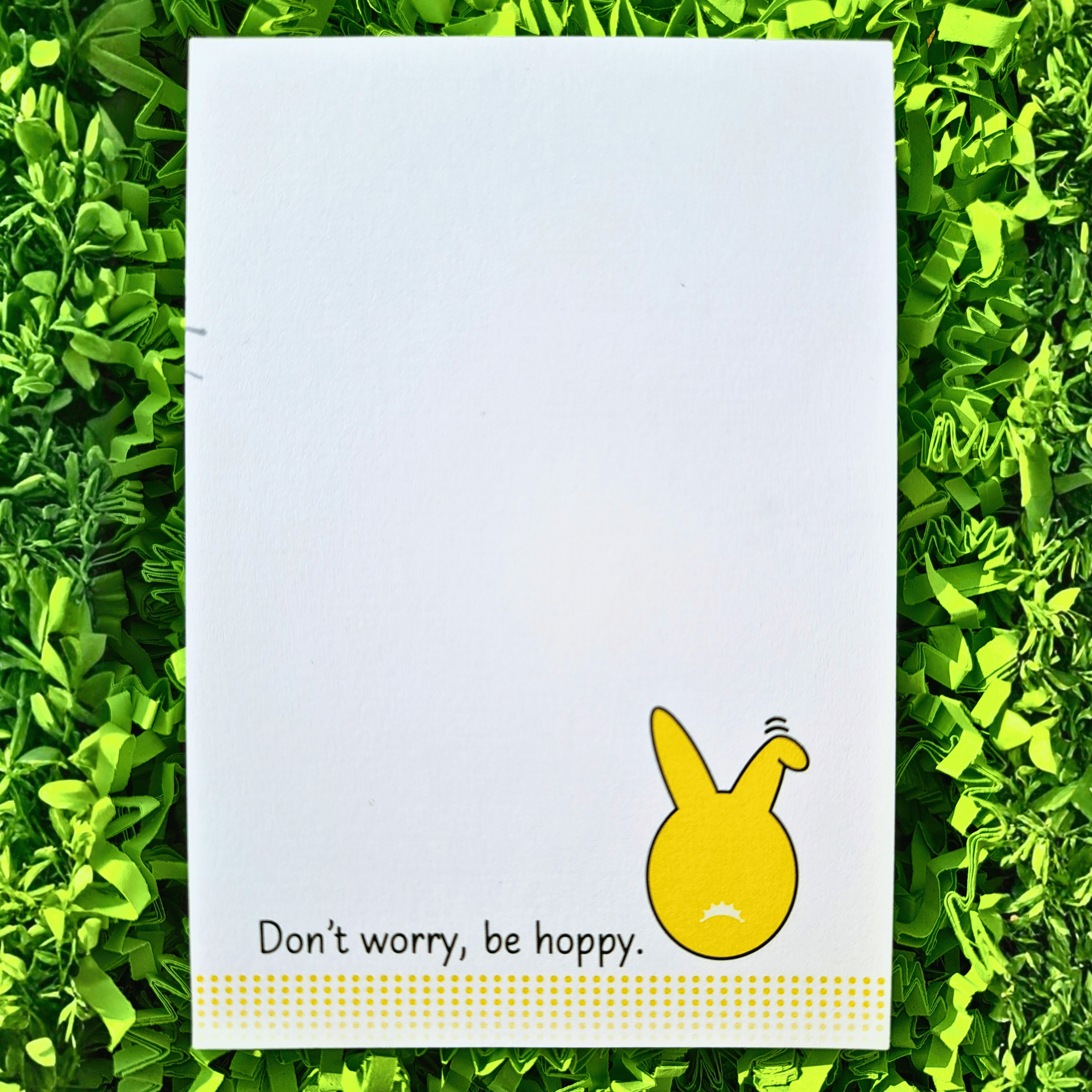 Notizblock Hase von Hinten. Spruch "Don't worry be hoppy". Frontalansicht Ein weißer DIN A7 Notizblock mit einem gelben Hasen-Popo-Motiv (Hase von hinten) unten rechts. Daneben steht der Spruch "Don’t worry, be hoppy.". Vier gepunktete Linien werden nach unten hin transparenter. Der Block liegt auf einem Hintergrund aus leuchte