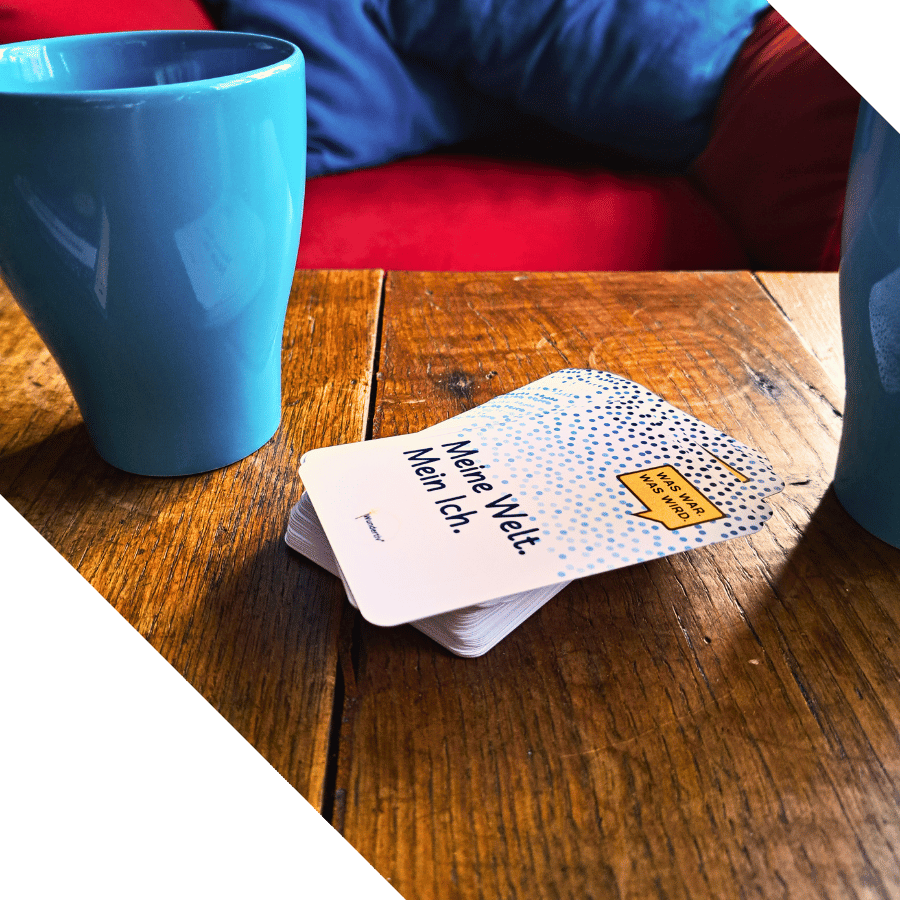 Gesprächswunder "Was war. Was wird. Meine Welt. Mein Ich". Dekorationsbild mit Tassen auf Holztisch. Schräg angeschnitten. Zwei türkis Tassen auf einem Holztisch. Zwischen den Tassen liegt ein Stapel Gesprächswunder-Karten "Was war. Was wird. Meine Welt. Mein ich.". Im Hintergrund ist ein zu roter Sessel mit türkis Kissen zu sehen. Das Bild ist oben und unten schräg angeschni