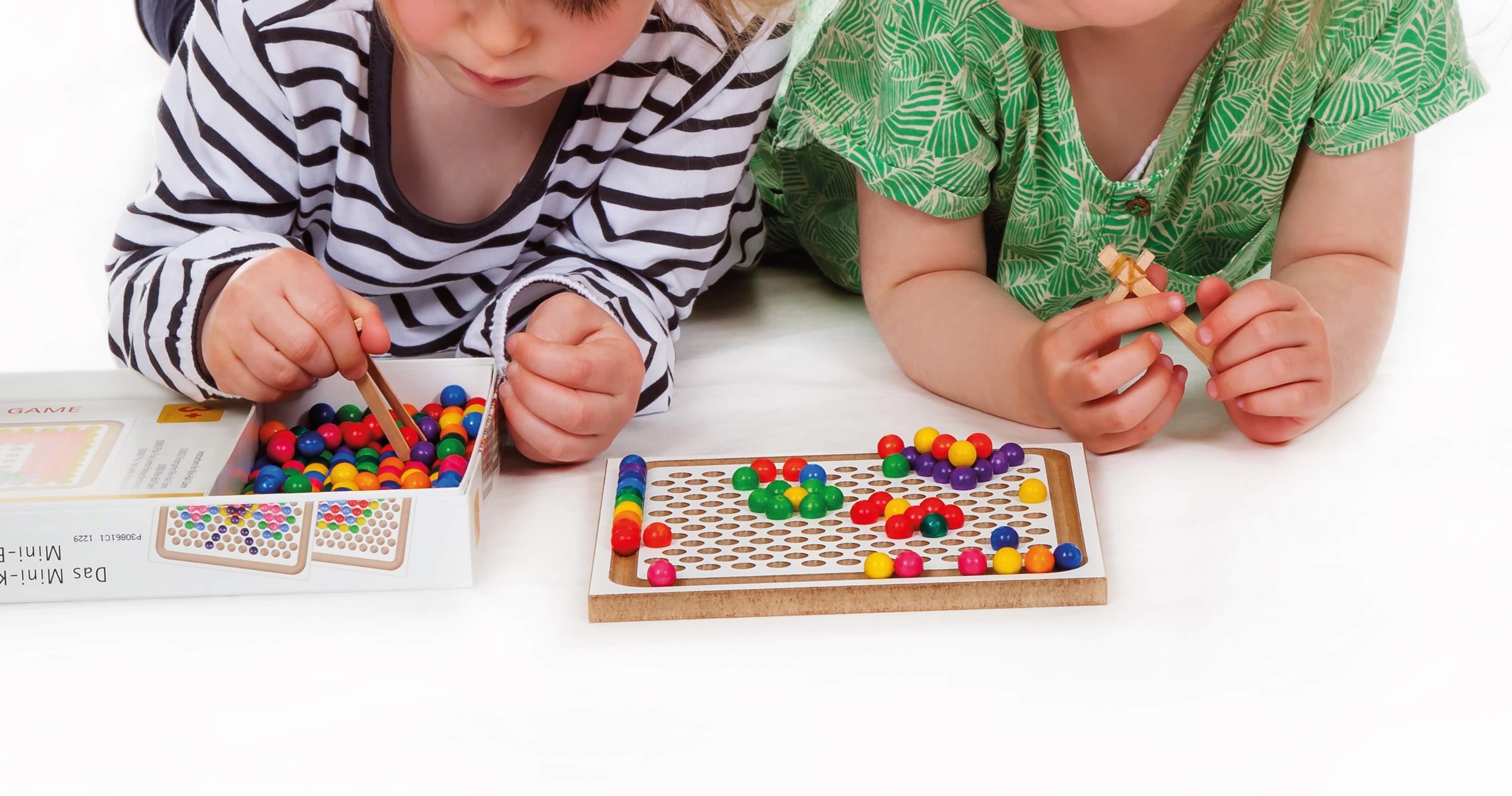 Zwei Kinder liegen auf einem weißen Boden und spielen mit dem Mini-Holzkugelspiel. Beide Kinder haben eine selbstgebastelte Pinzette in der Hand. Ein Kind nimmt mit dieser gerade eine Kugel aus der Verpackung auf. Auf dem Spielbrett sind bereits Kugeln vo