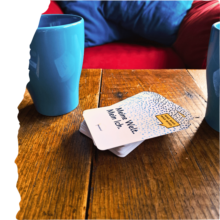 Gesprächswunder "Was war. Was wird. Meine Welt. Mein Ich". Dekorationsbild mit Tassen auf Holztisch. Links angerissen. Zwei türkis Tassen auf einem Holztisch. Zwischen den Tassen liegt ein Stapel Gesprächswunder-Karten "Was war. Was wird. Meine Welt. Mein ich.". Im Hintergrund ist ein zu roter Sessel mit türkis Kissen zu sehen. Bild ist links angerissen.