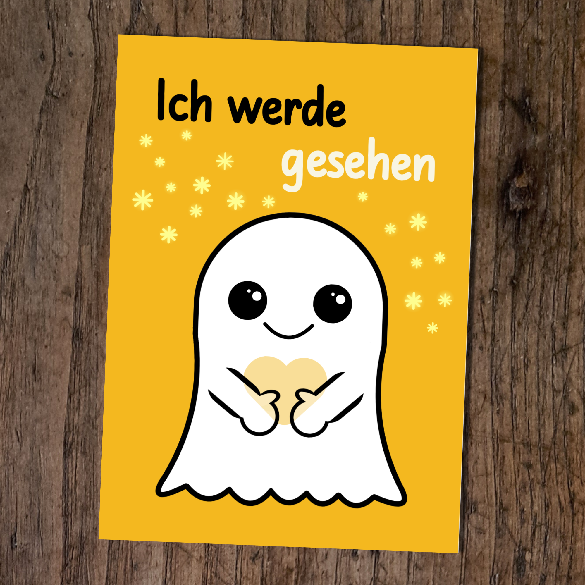 Gelbe-Orange Affirmations-Postkarte im Hochformat mit dem Spruch "Ich werde gesehen" und der Darstellung eines niedlichen Geistes, der ein Herz hält, liegend auf einem rustikalen Holztisch.